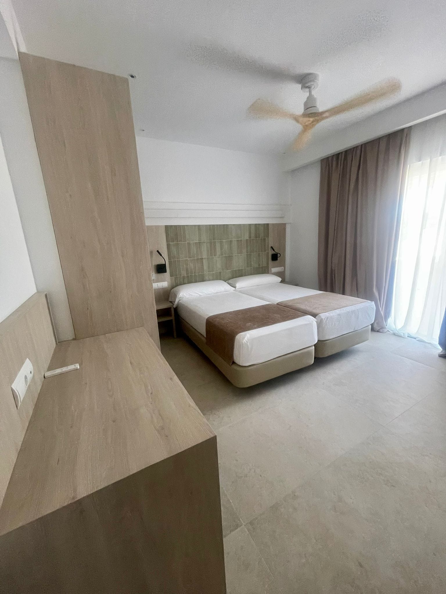Une chambre d’hôtel avec un lit, un bureau, un ventilateur de plafond et une fenêtre avec des rideaux.
