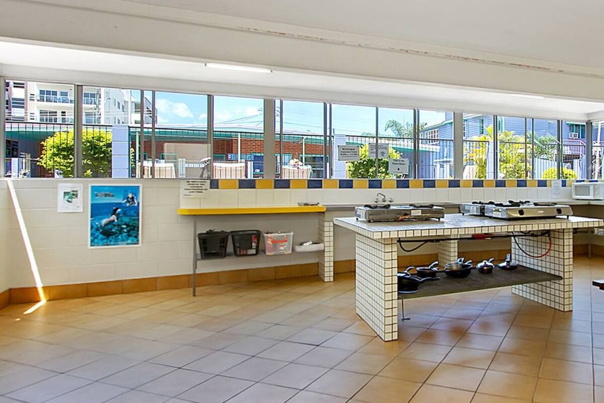 A kitchen with a tiled floor, a central island with cooking equipment, a wooden table, and large windows with a view of buildings and greenery.