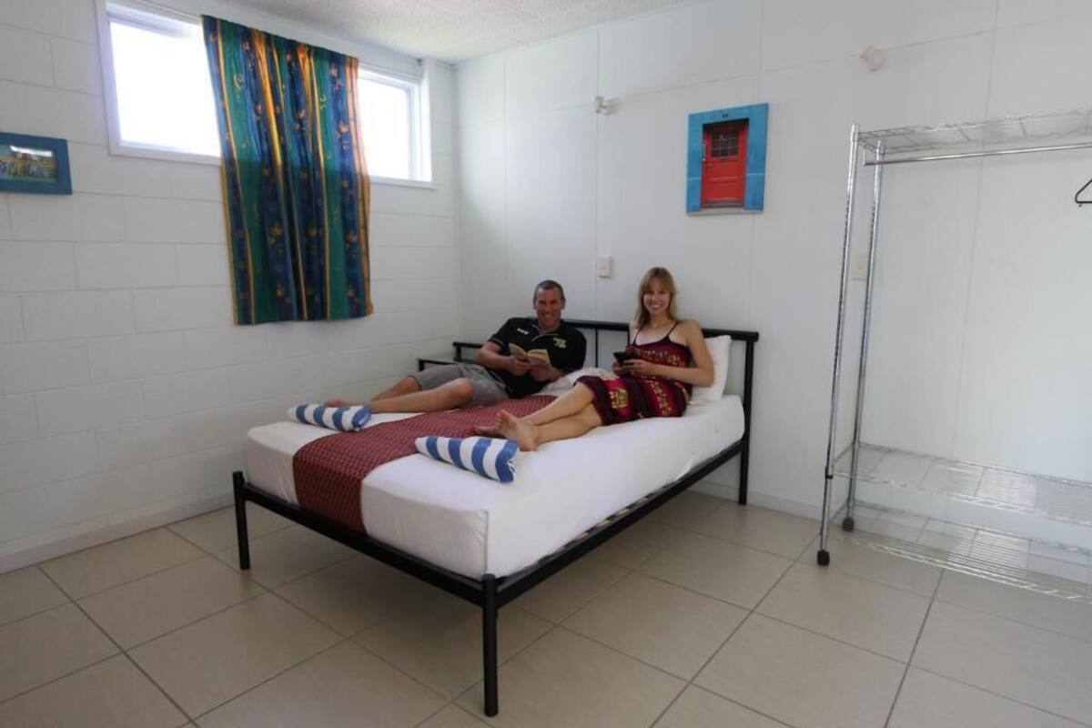 A single bed with a metal frame, white bedding, and a red blanket. There is a window with a colorful curtain and a small red-framed picture on the wall.