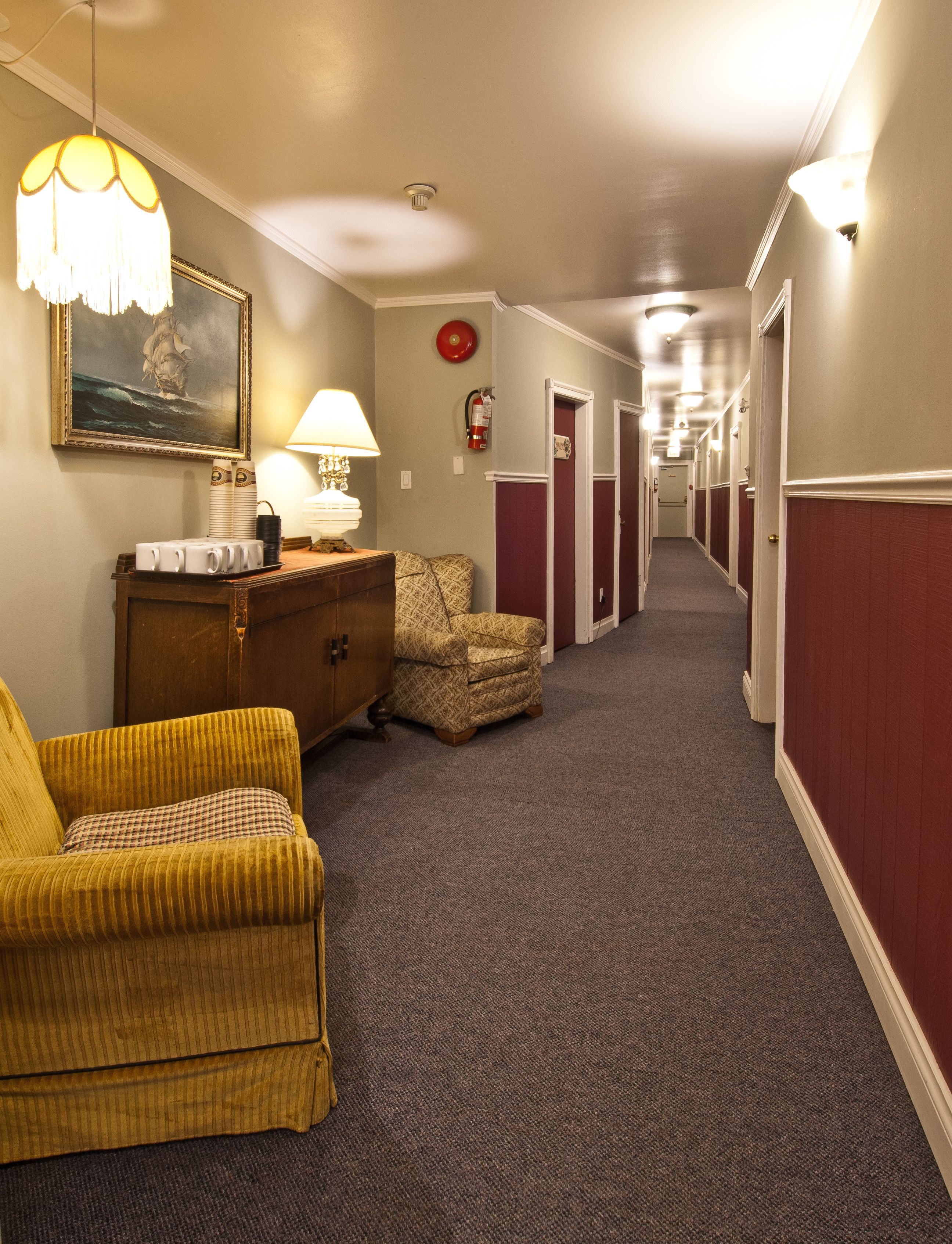 A hotel hallway with a patterned armchair, a wooden dresser, a lamp, and a framed picture on the wall.