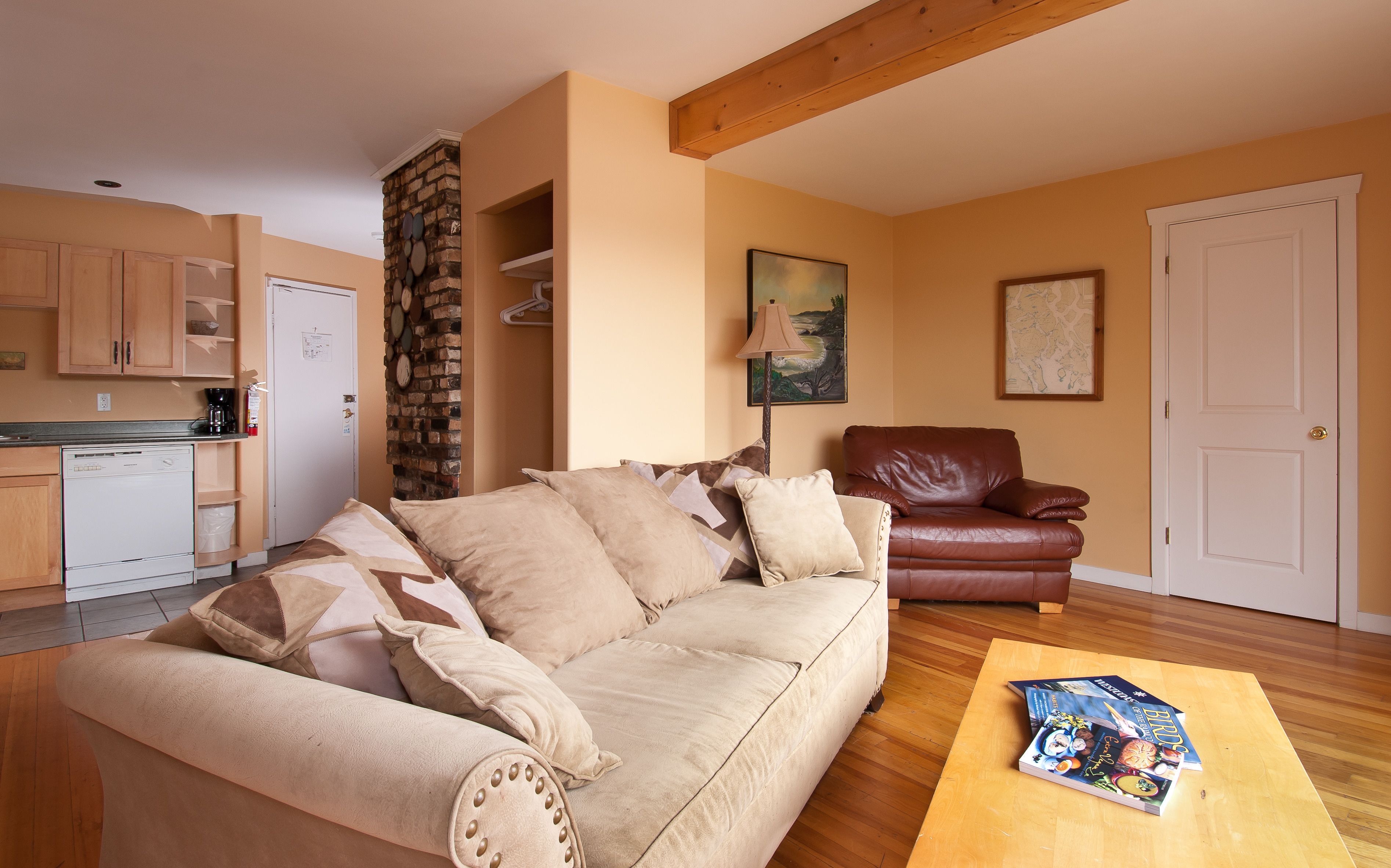 Un salon avec un canapé beige, un fauteuil en cuir marron et une table basse en bois. Au fond, on aperçoit une cuisine avec des meubles en bois et un réfrigérateur blanc.