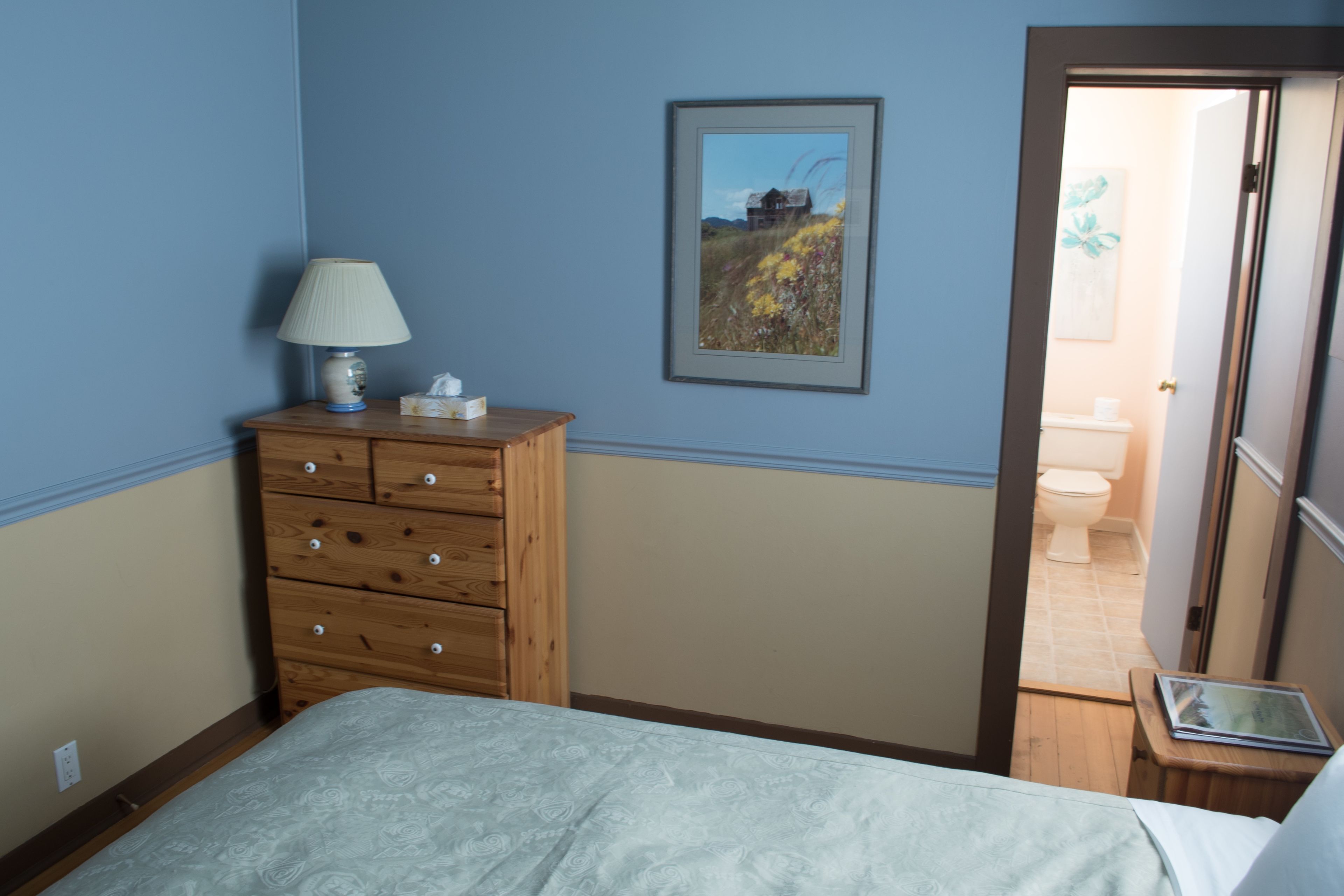 Un dormitorio con cama, cómoda, lámpara, un cuadro enmarcado y vista al baño.