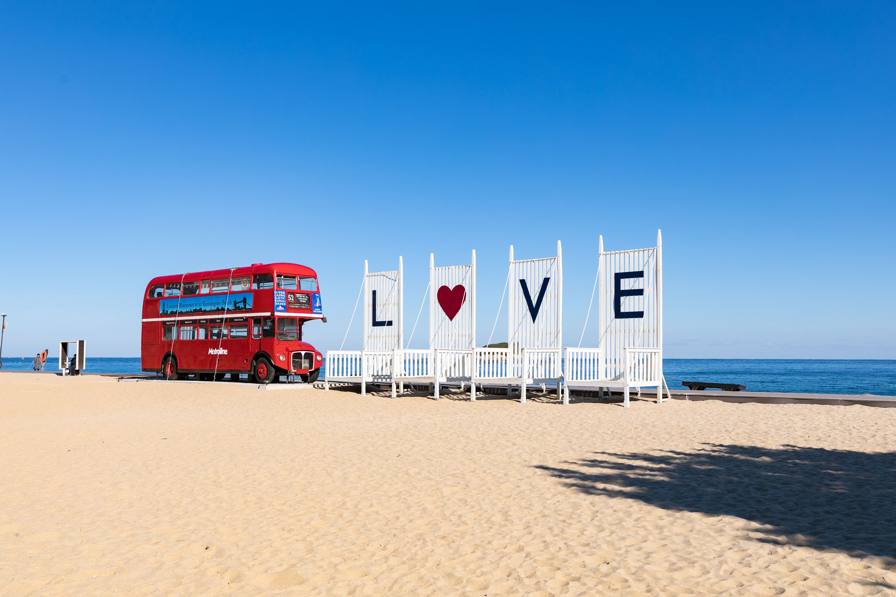 Un autobus à deux étages rouge est garé sur une plage de sable, et une clôture blanche affiche les mots « LOVE » et un symbole en forme de cœur.