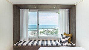 Een slaapkamer met een groot raam dat uitkijkt op een strand, een bed met gestreepte lakens en een bank met kussens.