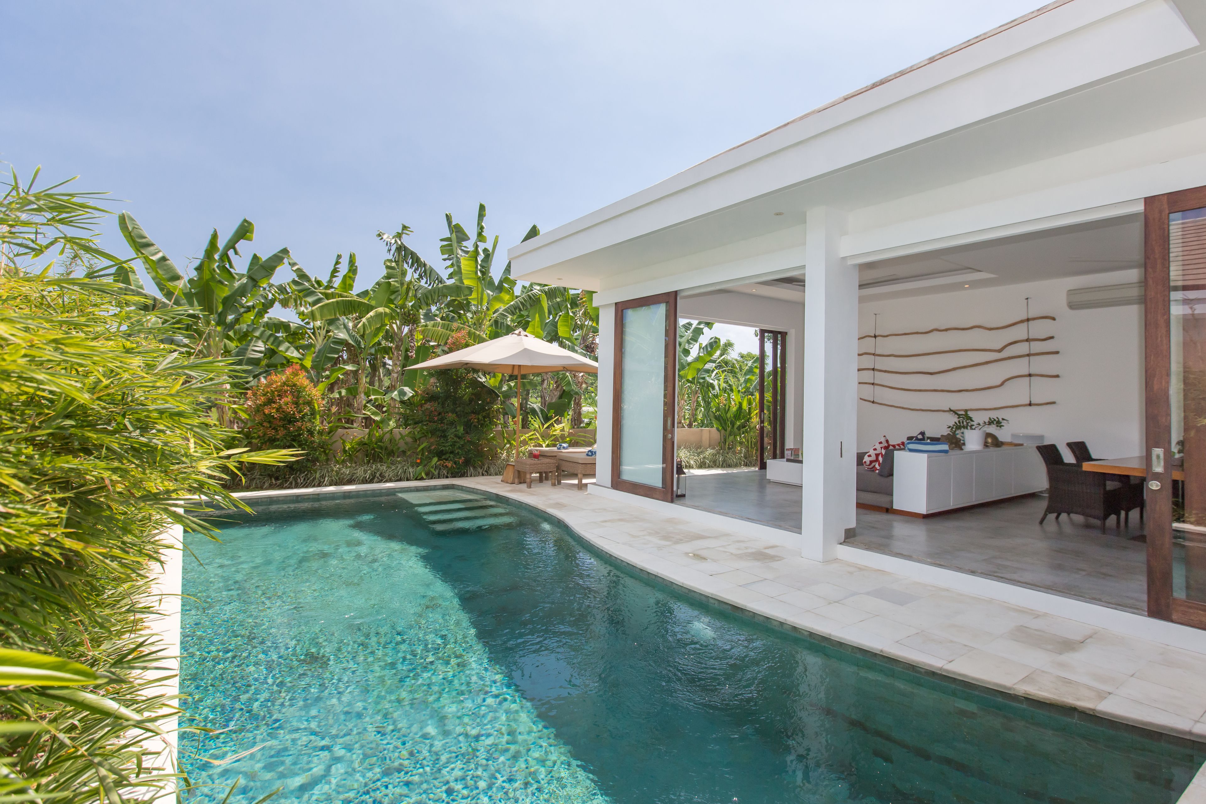 A modern house with a swimming pool, a patio with a table and chairs, and a glass door leading to an indoor area with a sofa and a dining table.