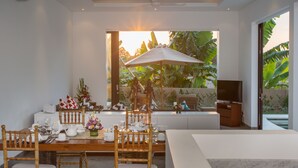 A dining area with a wooden table set for four, surrounded by bamboo chairs. A large umbrella provides shade, and the room features a large window with a view of lush greenery outside.