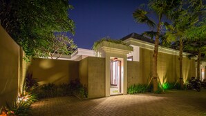 A paved entrance to a building with a lit doorway, surrounded by palm trees and plants.