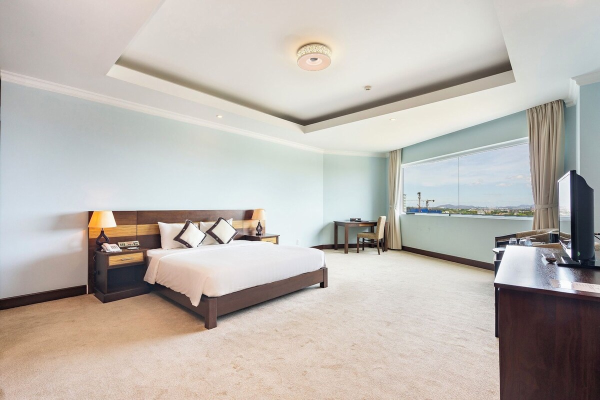 A hotel room with a bed, bedside tables, a desk, a chair, a TV, and a large window with a view.