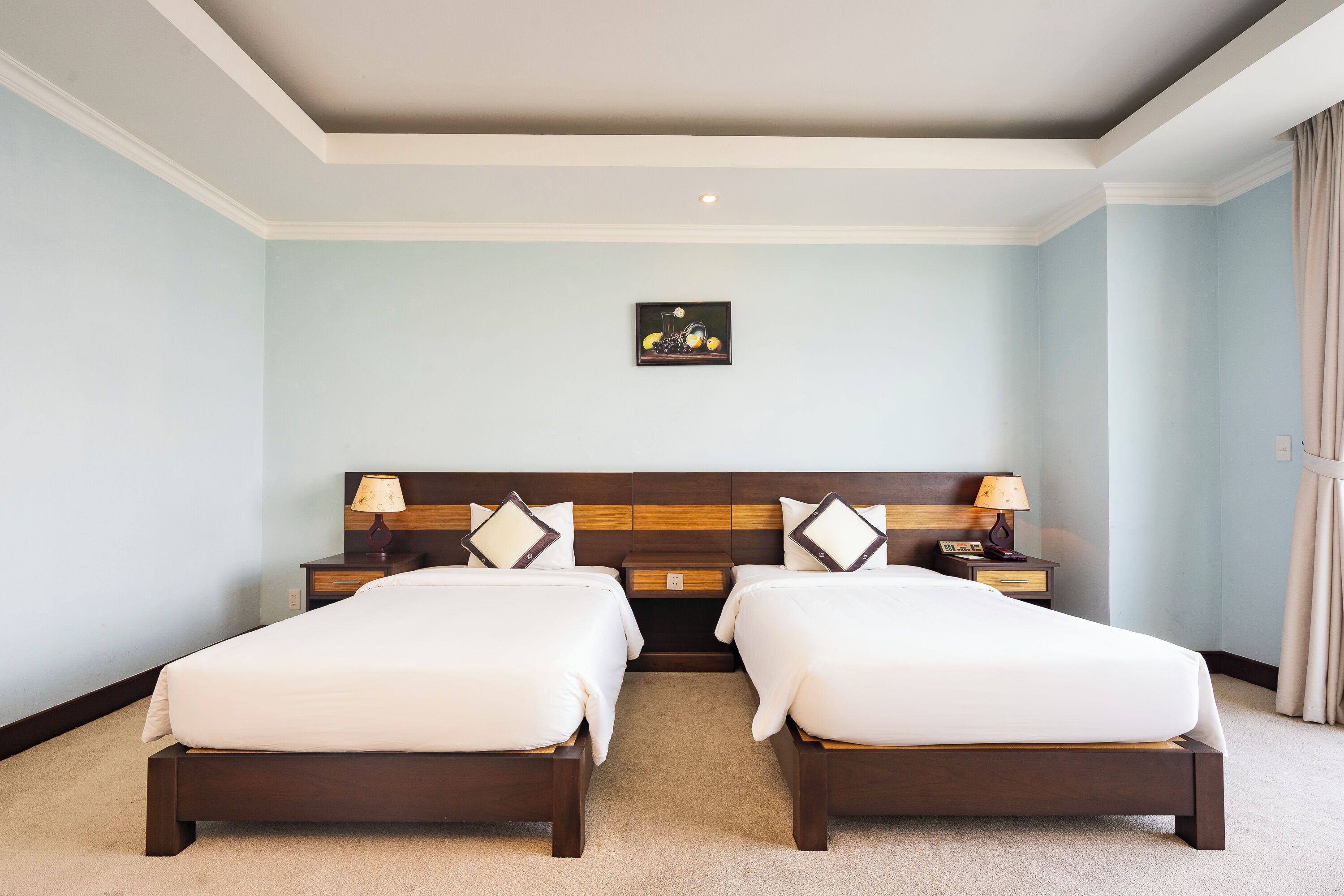 A hotel room with two beds, a wooden headboard, bedside tables with lamps, a small artwork on the wall, and a beige carpet.