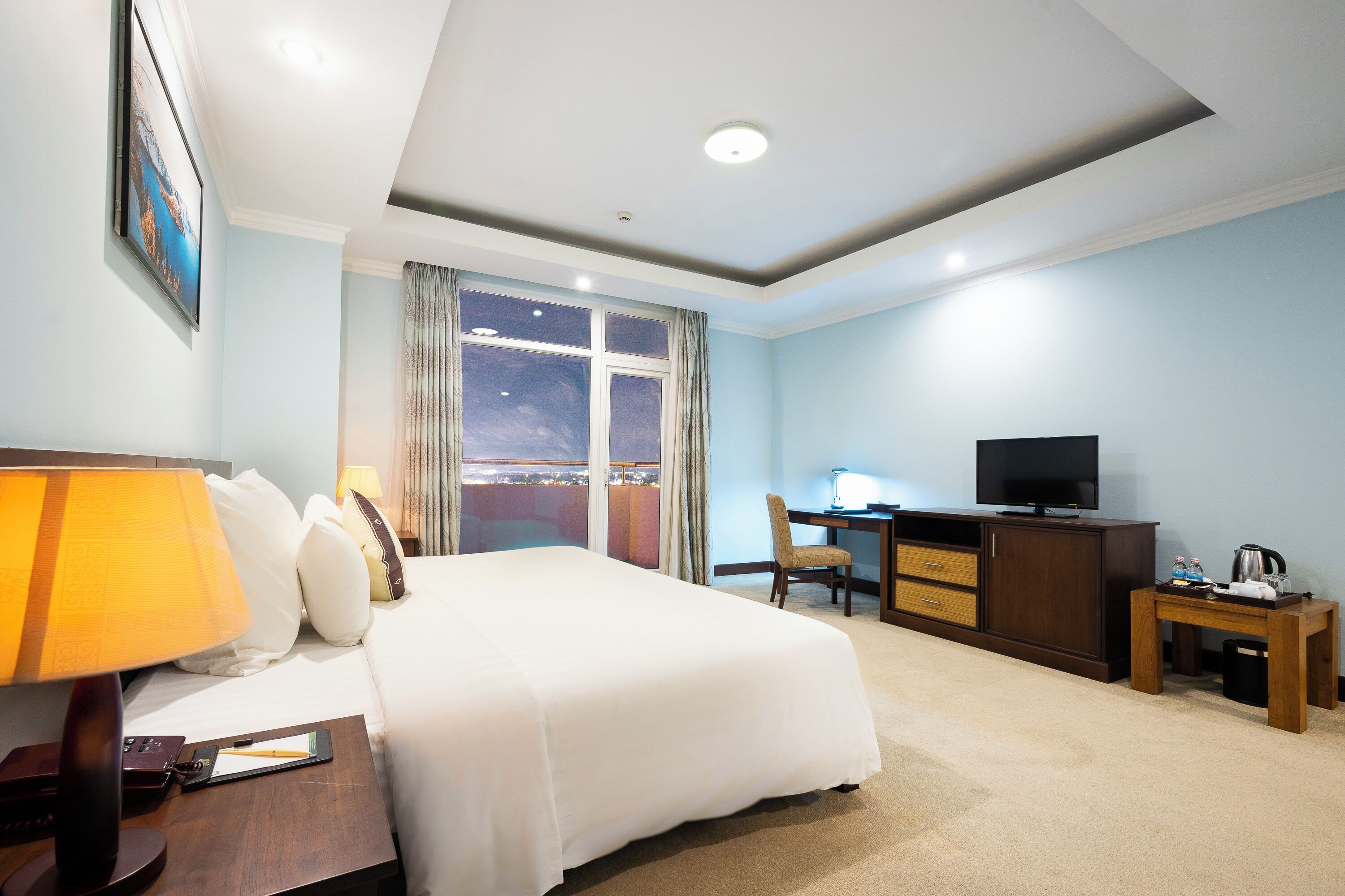 A hotel room with a bed, desk, television, and a view of the city.