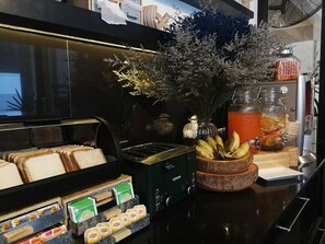 A kitchen counter with a bread display, a fruit bowl, a coffee machine, and a juice dispenser. - 3Howw Hostel at Sukhumvit 21 (Bangkok)