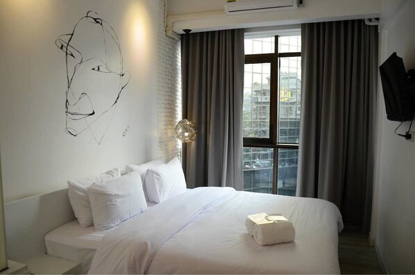 A modern hotel room with a large bed, white bedding, a wall-mounted abstract art piece, a large window with curtains, and a view of the cityscape. - 3Howw Hostel at Sukhumvit 21 (Bangkok)
