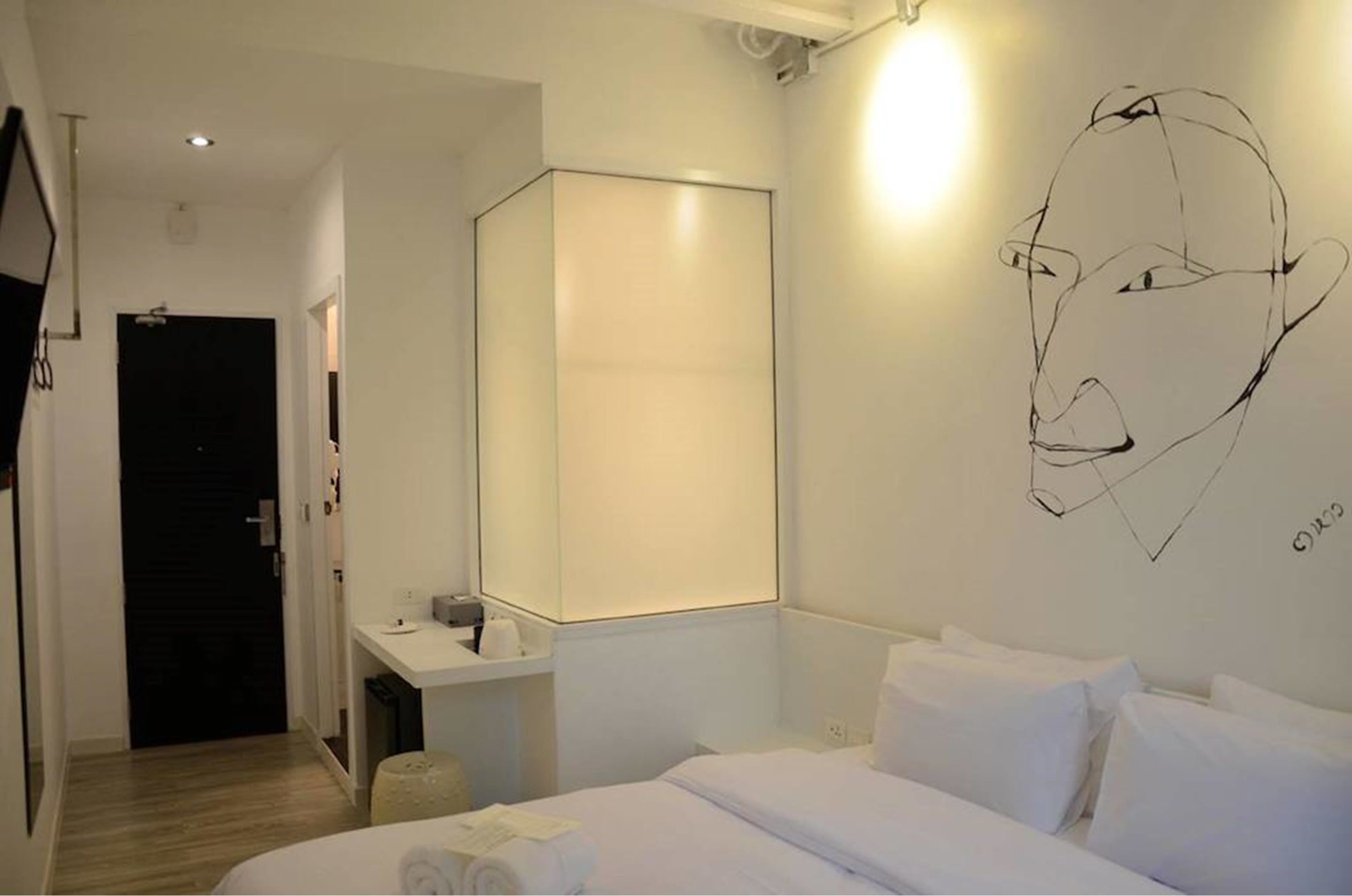 A hotel room with a bed, a bedside table, a mirror, and a wall with a simple line drawing.