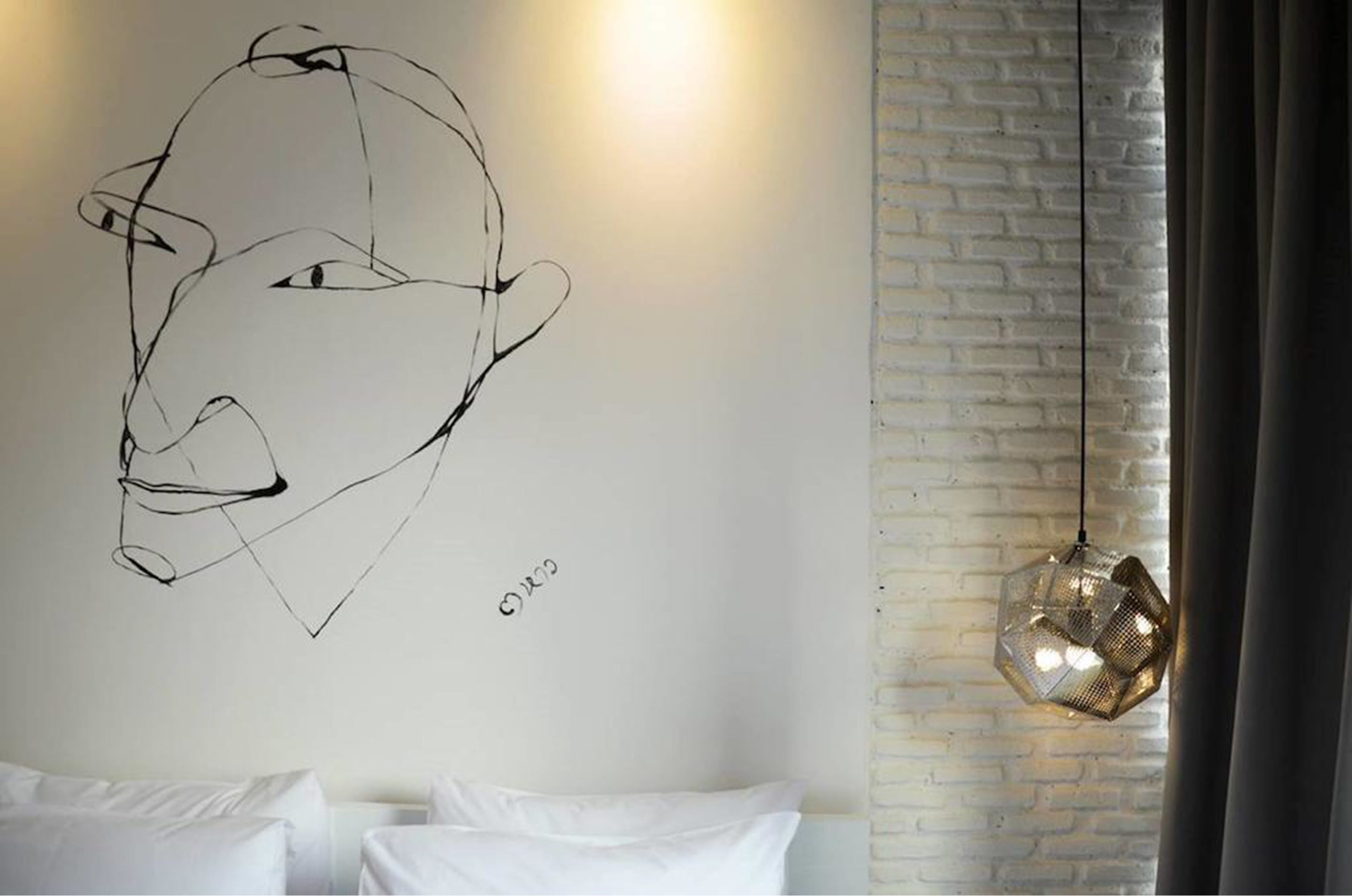 Une pièce avec un mur en briques blanches, un luminaire suspendu aux formes géométriques et un lit blanc avec des coussins.