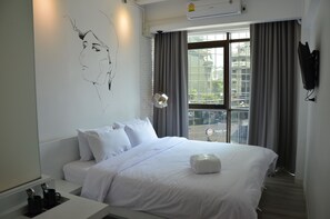 A modern hotel room with a large bed, a wall art piece, a desk with a lamp, and a view of the city through large windows. - 3Howw Hostel at Sukhumvit 21 (Bangkok)