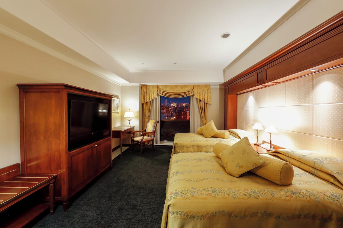 Habitación de hotel con dos camas, televisión, escritorio y vista a un paisaje urbano nocturno.