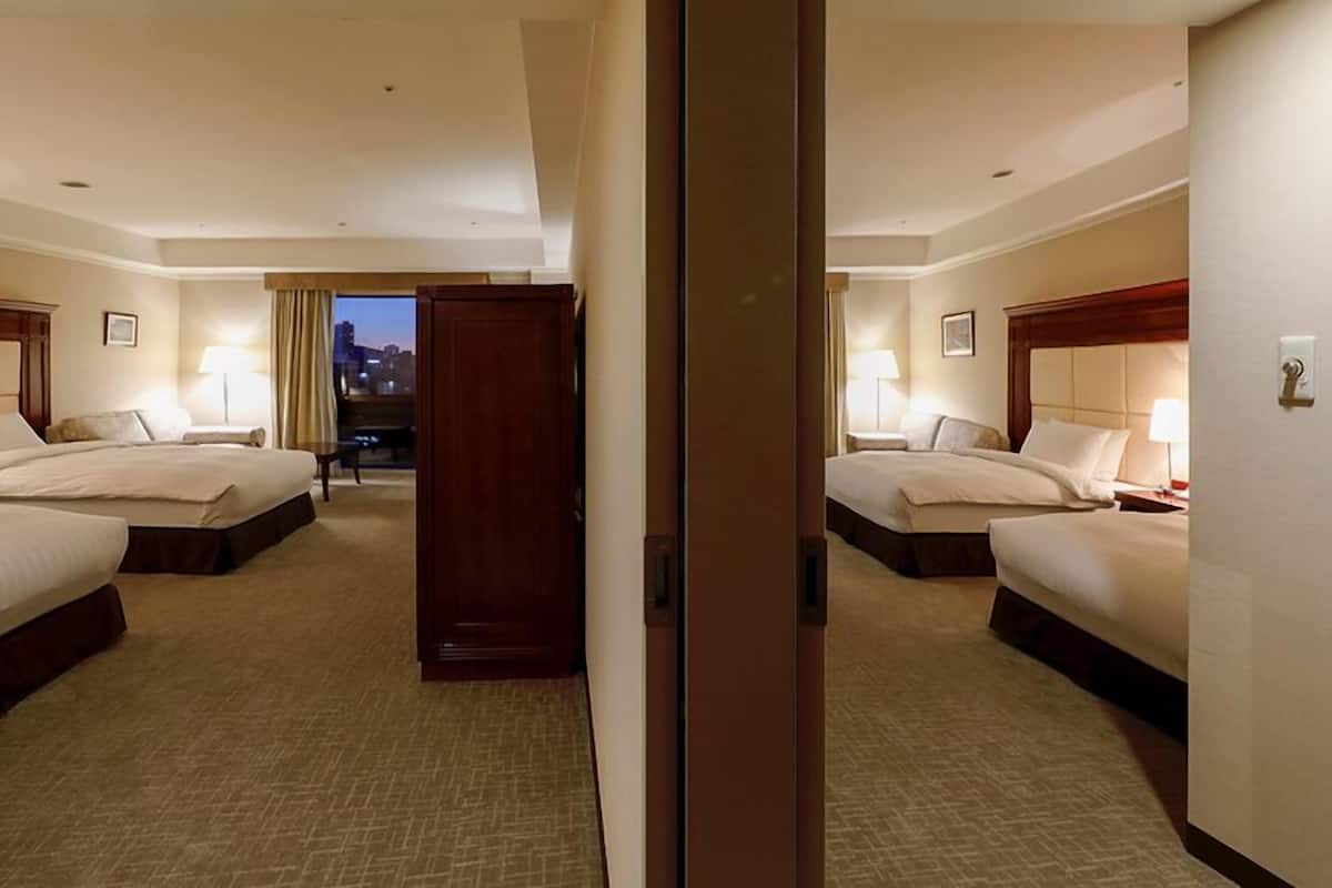 Habitación de hotel con dos camas, una mesita de noche con lámpara, vista a la ciudad al anochecer y una puerta de madera.