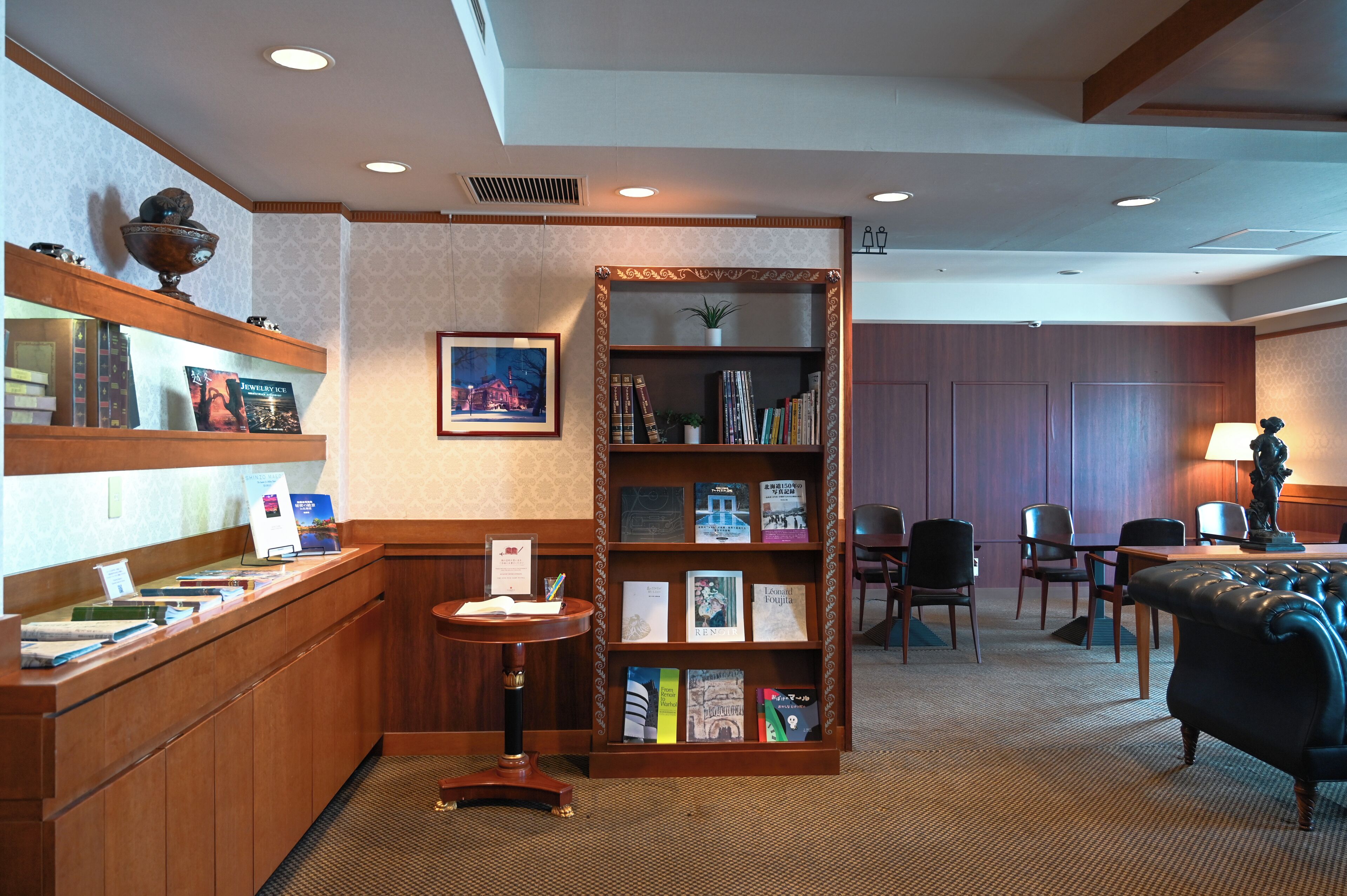 Um hall de hotel com um balcão de recepção de madeira, uma estante com livros e um quadro emoldurado, uma mesa redonda com cadeiras e uma poltrona confortável.