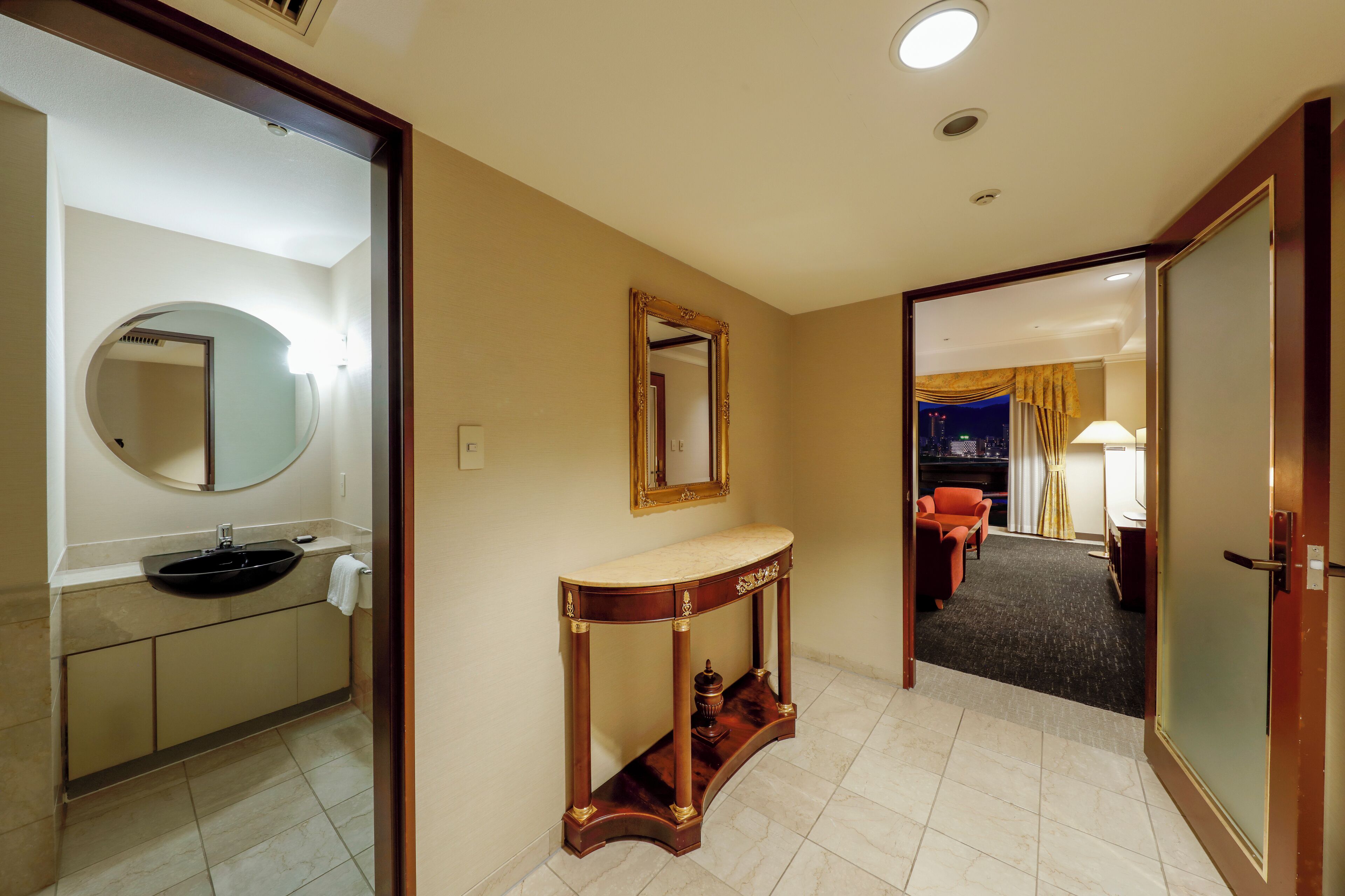 A hotel room with a bathroom featuring a sink, mirror, and vanity. There is a console table with a lamp and a view of the city at night.