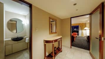 A hotel room with a bathroom featuring a sink, mirror, and vanity. There is a console table with a lamp and a view of the city at night.