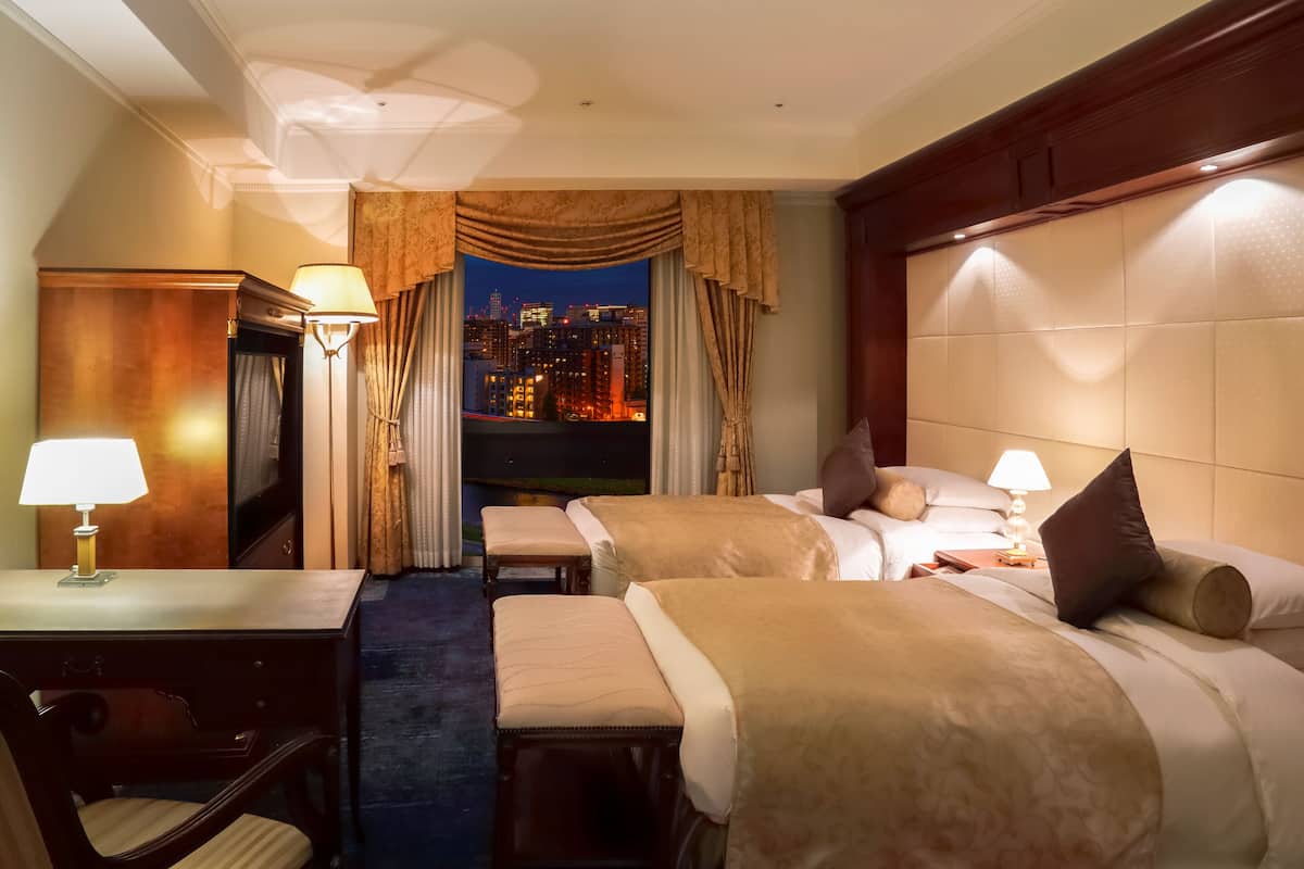 Habitación de hotel con dos camas, un escritorio y una silla. Se ve un paisaje urbano a través de la ventana.