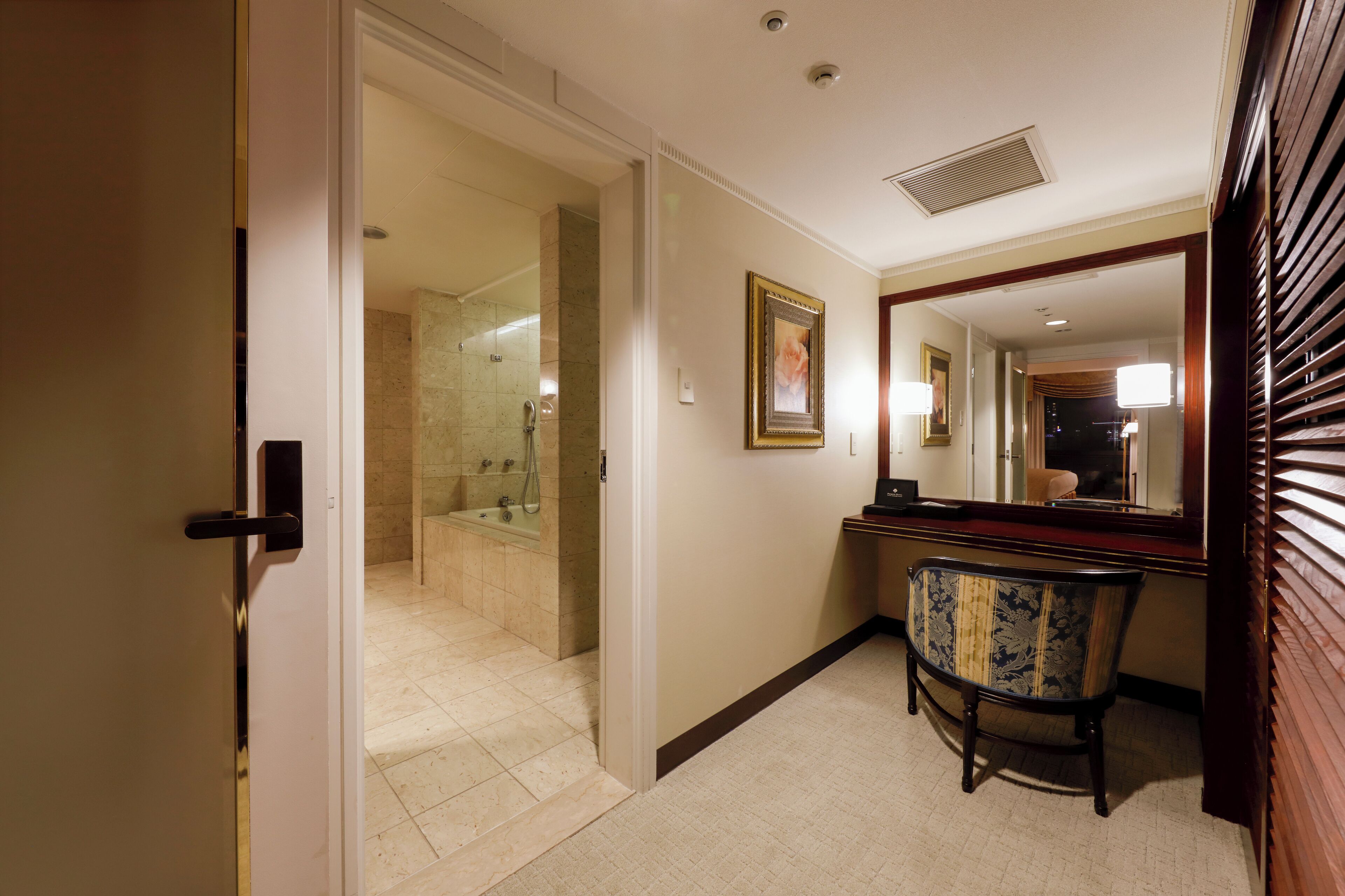 A hotel hallway with a bathroom, a chair, and a mirror.