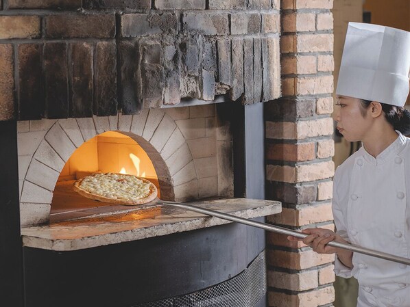 A brick oven pizza oven with a pizza inside, a chef in a white uniform, and a pizza peel in front of it. - Premier Hotel Tsubaki Sapporo (Sapporo)