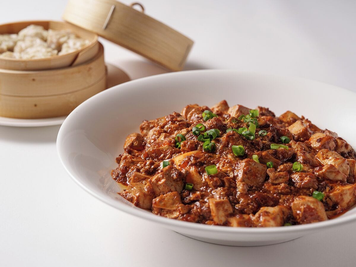 A bowl of spicy meat dish garnished with green onions, accompanied by a bamboo steamer in the background.