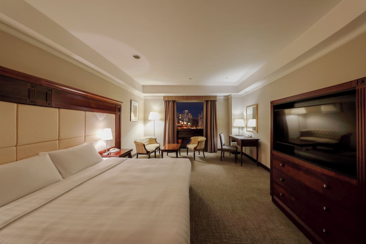 Habitación de hotel con una cama grande, un televisor, un escritorio con lámpara, dos sillones y vistas a un paisaje urbano nocturno.