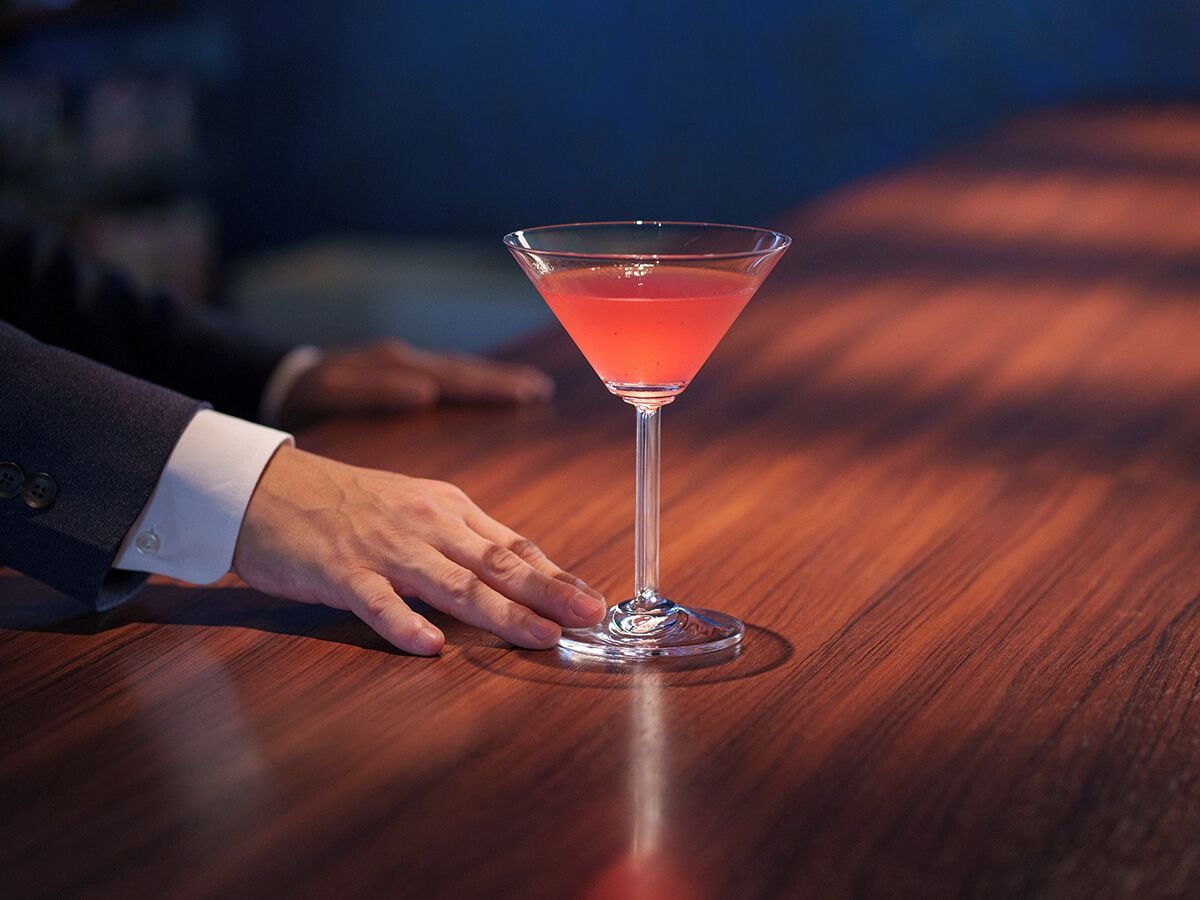 A martini glass on a wooden table with a hand reaching towards it.