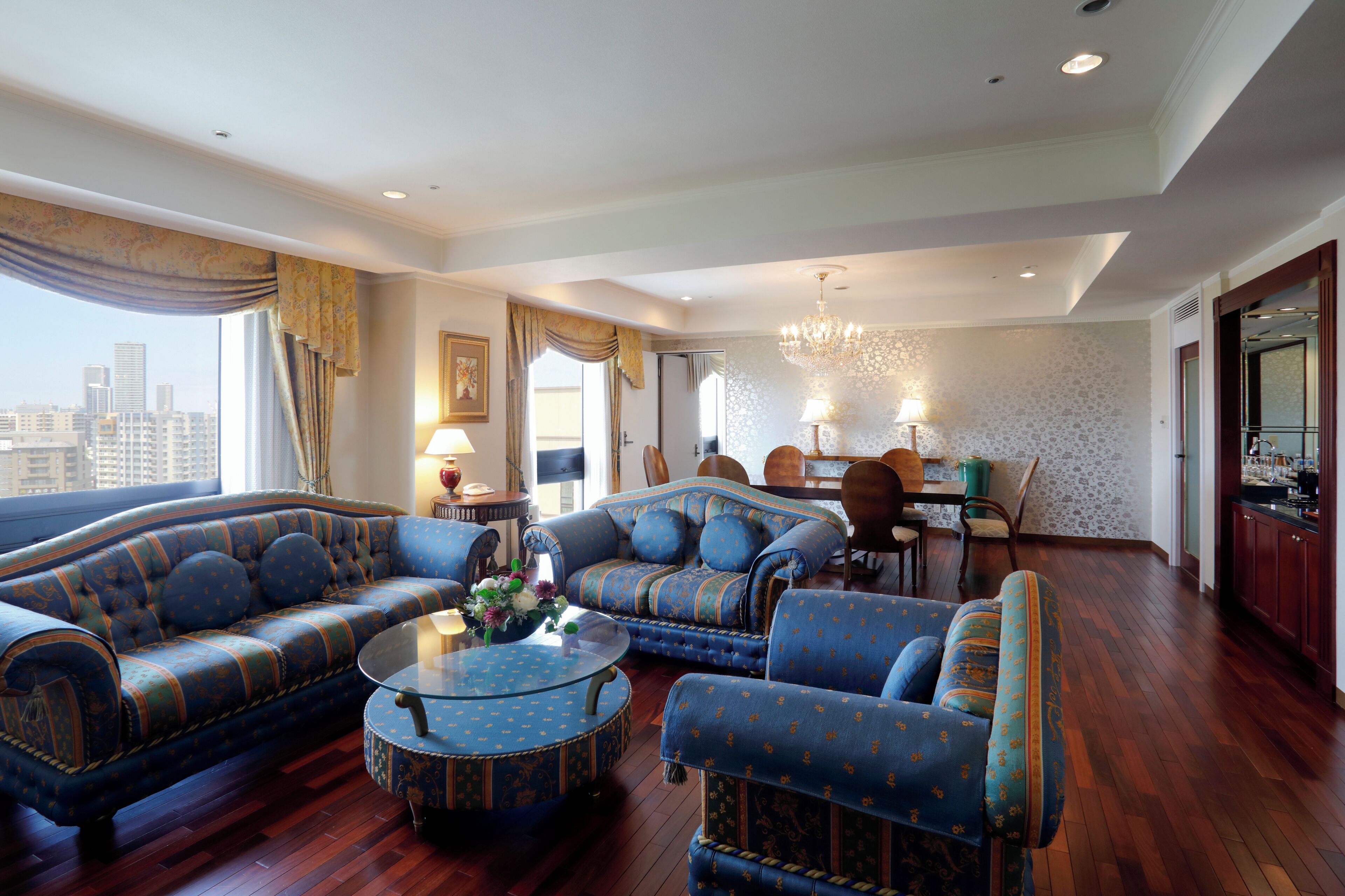 A spacious living room with a large sofa, a smaller sofa, and two armchairs. There is a dining area with a table and chairs, and a kitchen area with cabinets and a countertop.