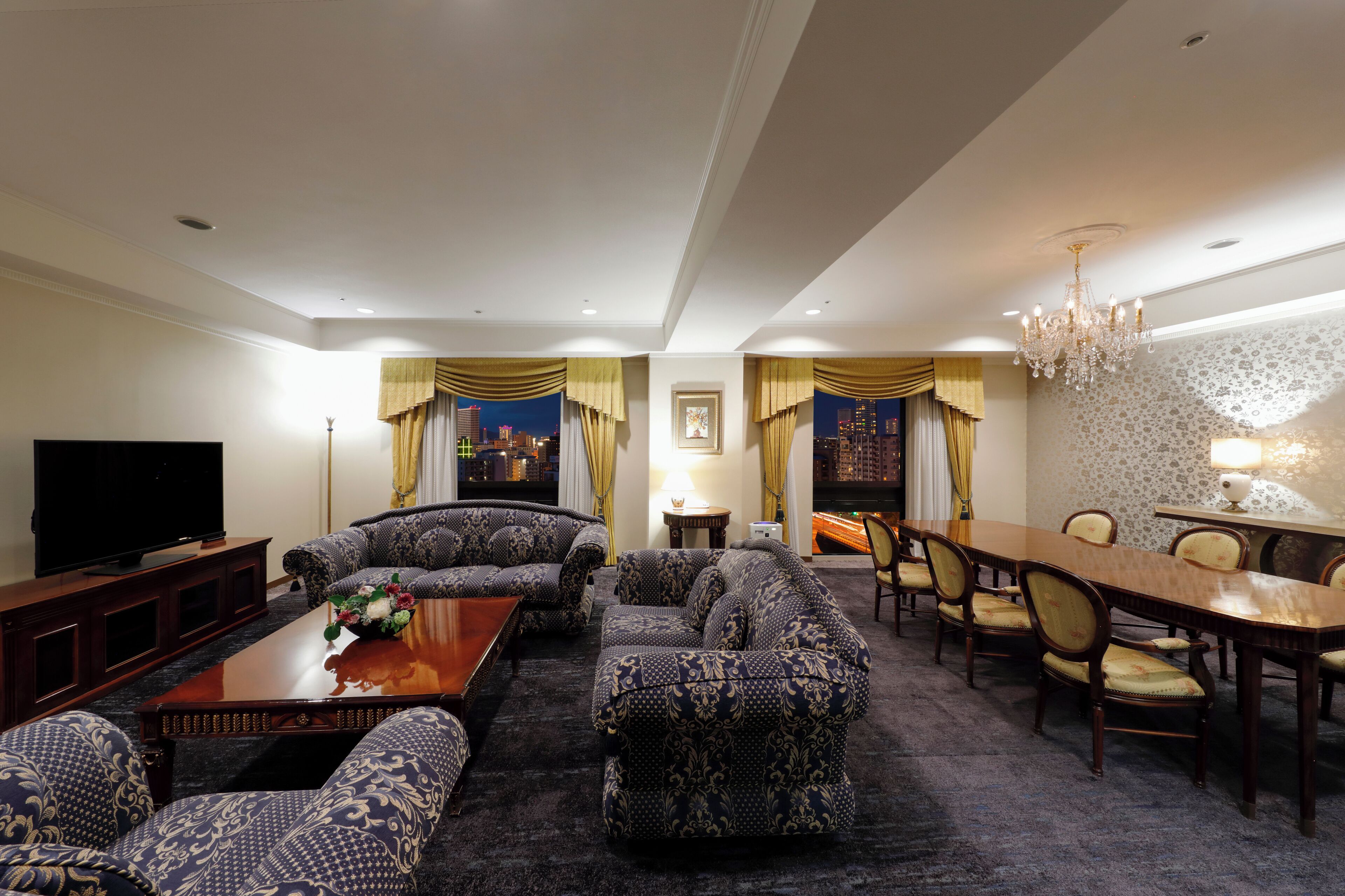 A spacious living room with a large sofa set, a dining table with chairs, a flat-screen TV, and a chandelier.