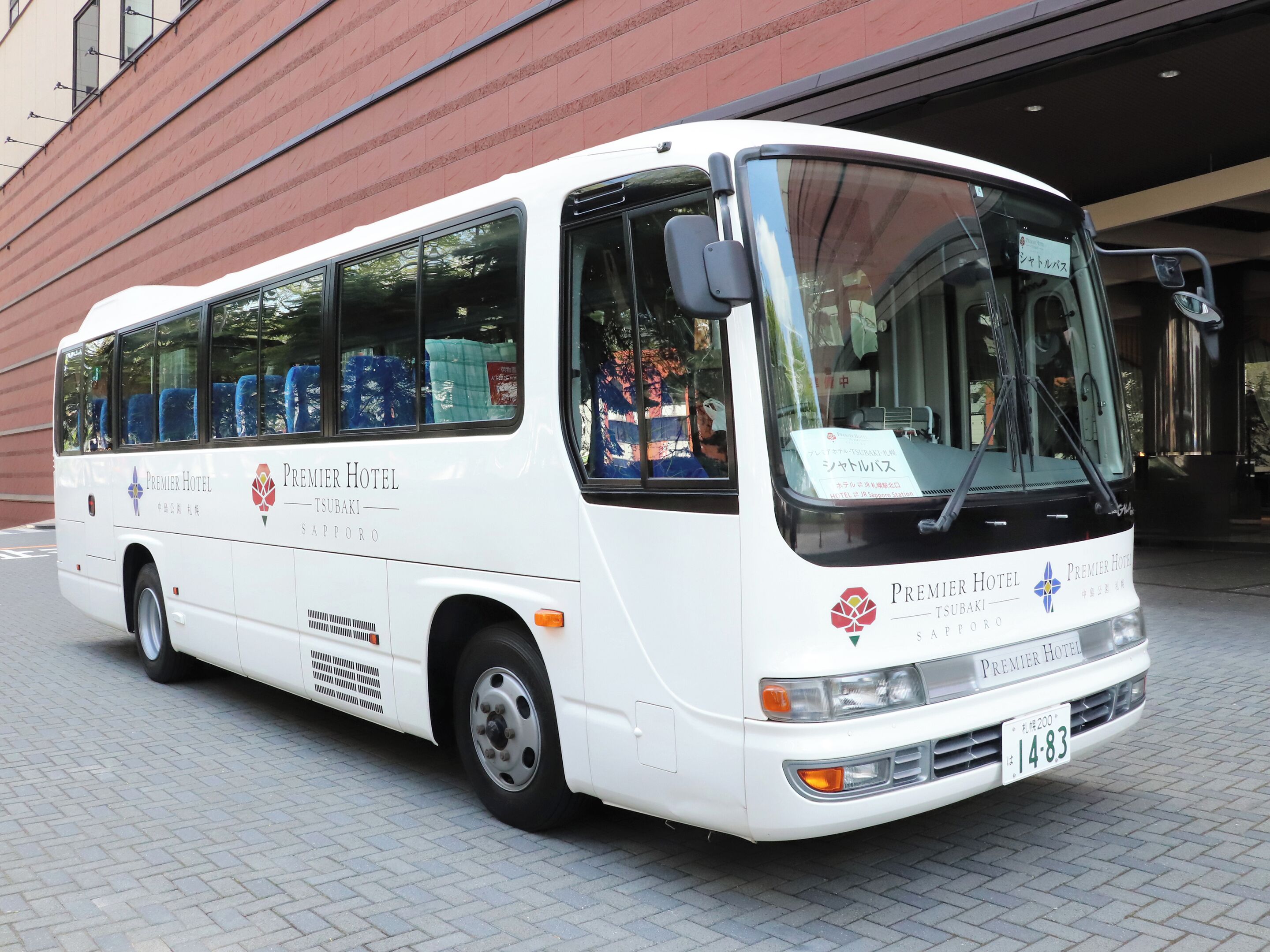 A white bus with "Premier Hotel Tsukaki Sapporo" branding, parked in front of a building.