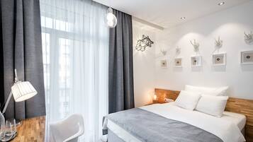 Een hotelkamer met een groot bed, een bureau met een lamp, en een stoel. Gordijnen, een wandlamp en lijstjes met foto's sieren de wand.