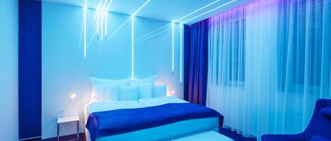 A modern hotel room with a large bed, a bedside table, a chair, and a view of the city through sheer curtains.