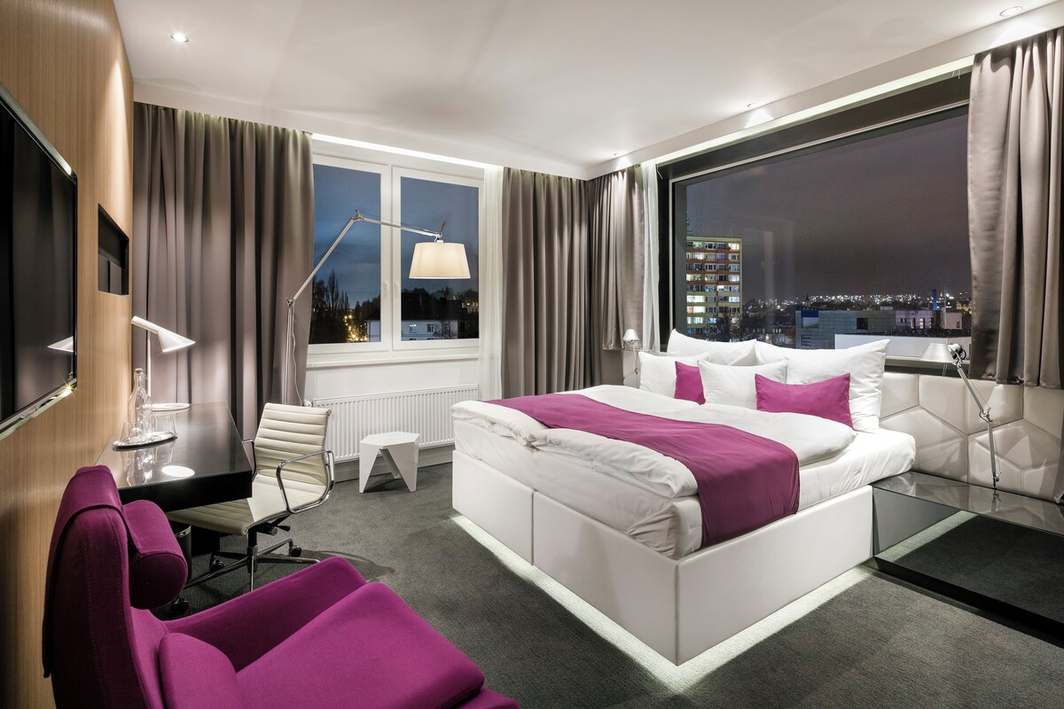 Habitación de hotel moderna con una cama grande, un escritorio, una silla y vistas de la ciudad por la noche.