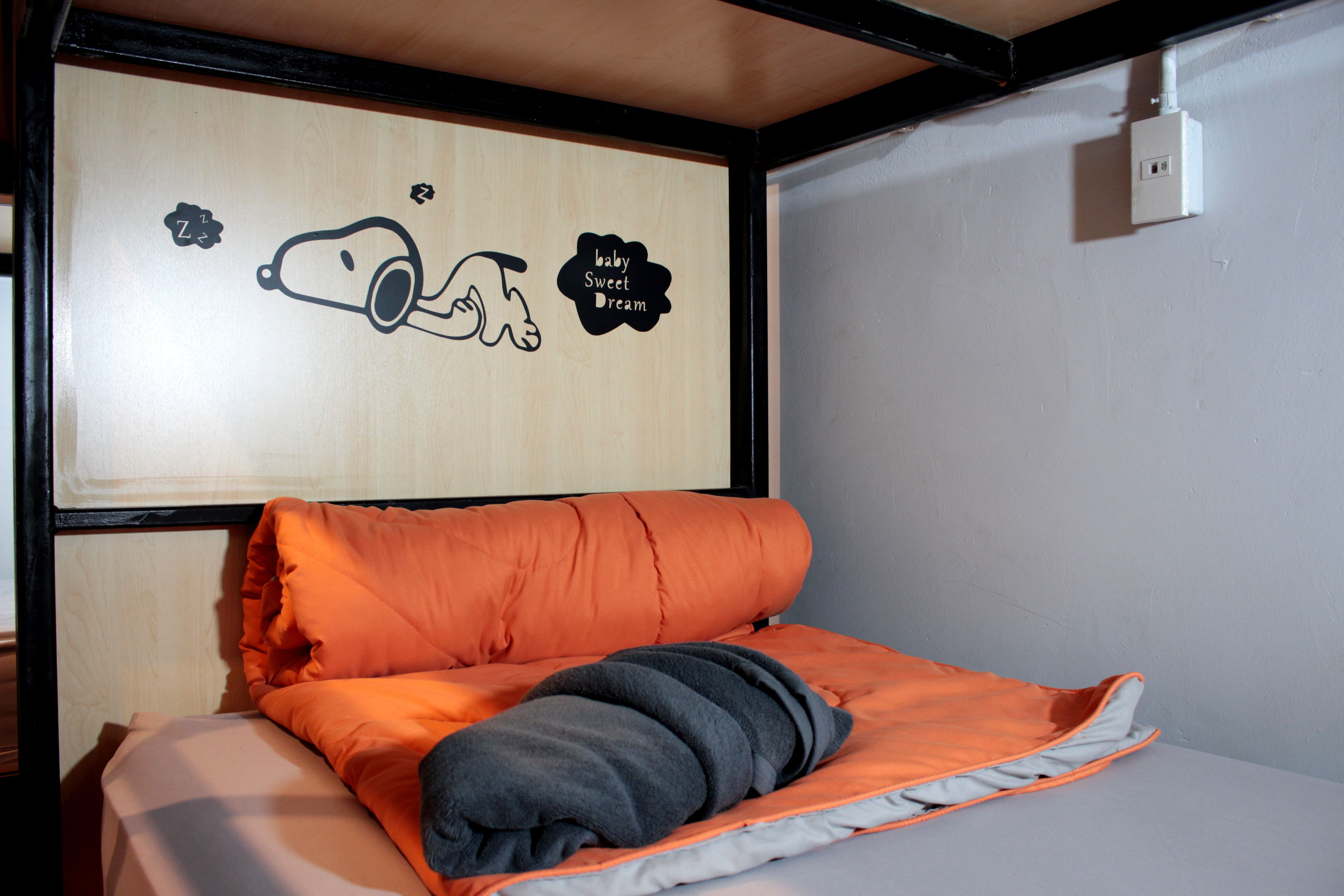 A bed with an orange blanket and a grey pillow, a wall with a cartoon drawing, and a power outlet on the wall.