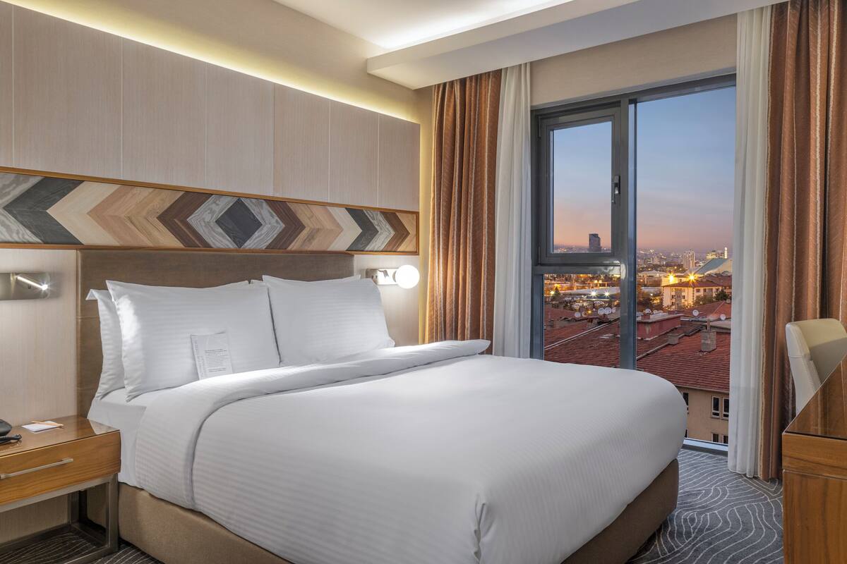 A hotel room with a large bed, a bedside table with a phone, a flat-screen TV, and a city view through the window.