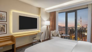 A hotel room with a large bed, a desk with a chair, a TV, and a view of a cityscape with a prominent building. - Mercure Ankara Kızılay (Ankara)