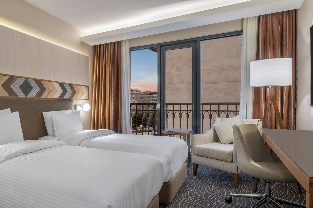 A hotel room with two beds, a desk, a chair, a lamp, and a view of the city.