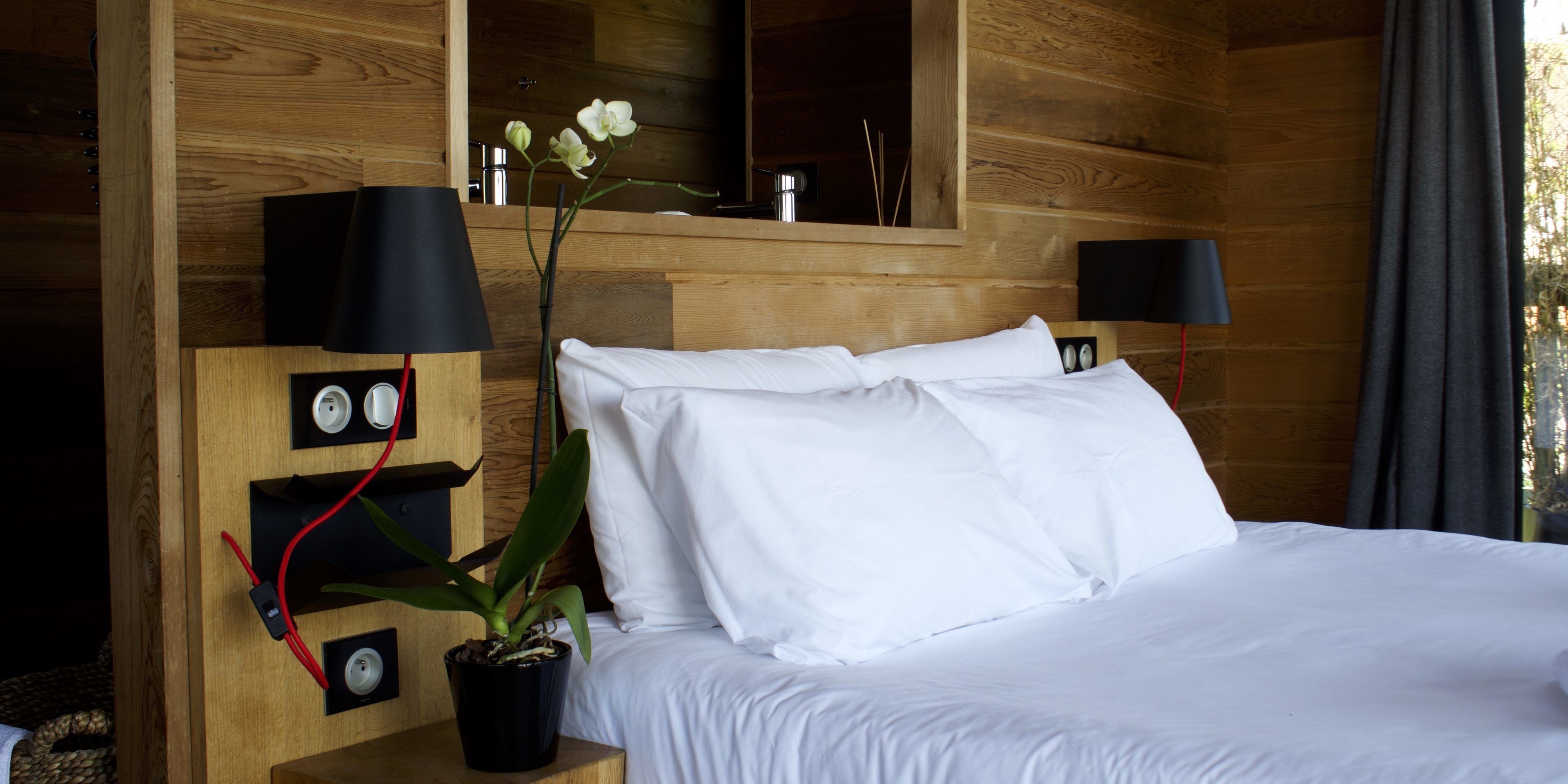 A wooden room with a bed, two wall-mounted lamps, a potted plant, and a mirror.