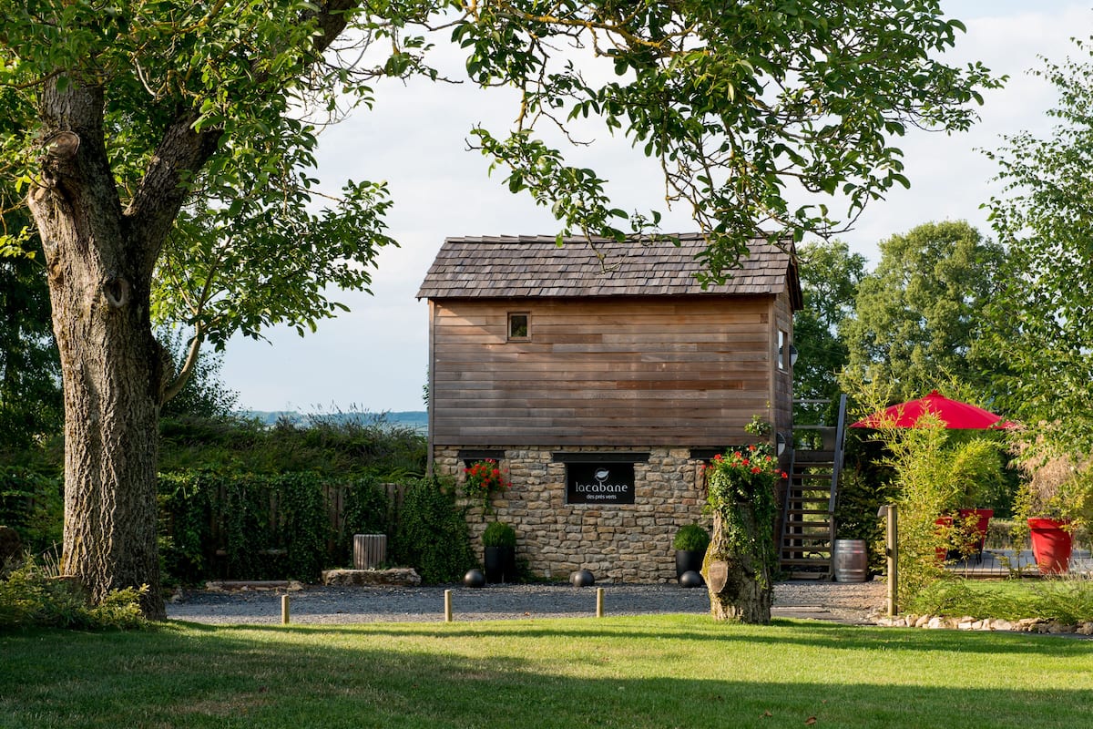 Un bâtiment en bois avec une façade en pierre, une enseigne indiquant « la cabane des pets verts », et un parapluie rouge en arrière-plan.