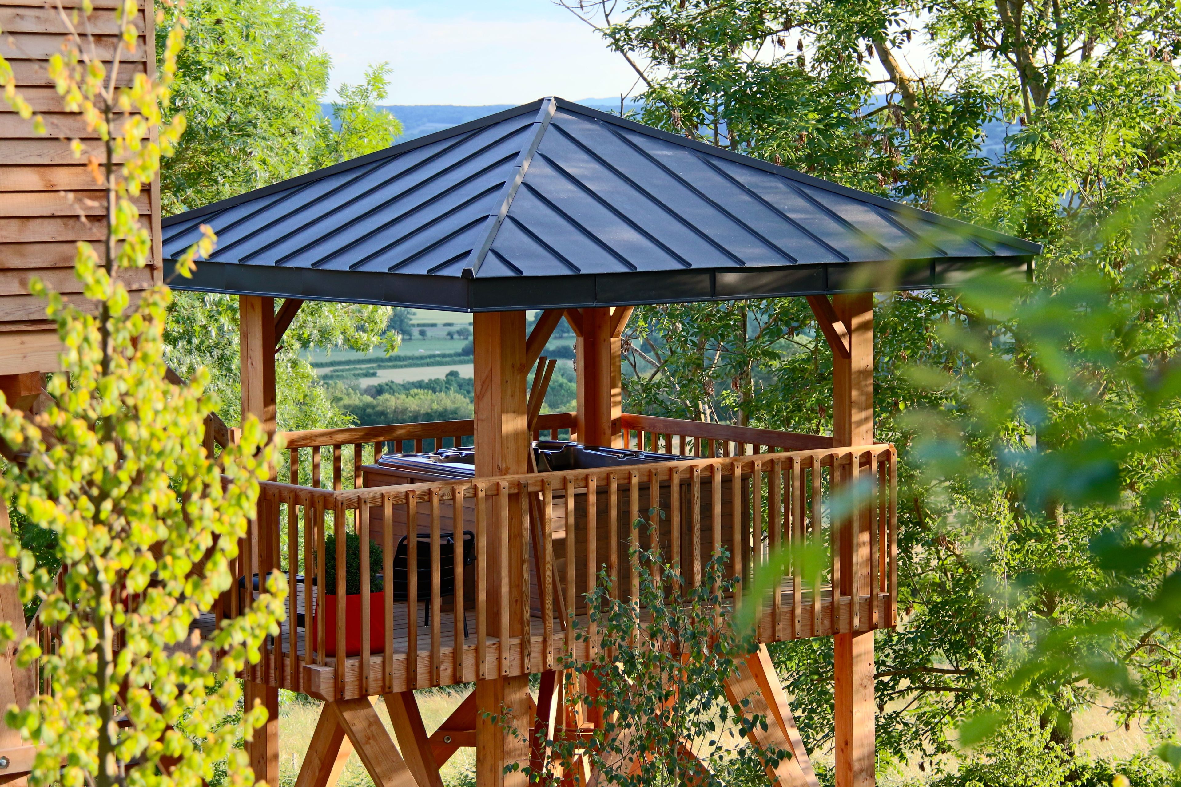 A wooden gazebo with a metal roof, surrounded by trees and a view of the countryside.