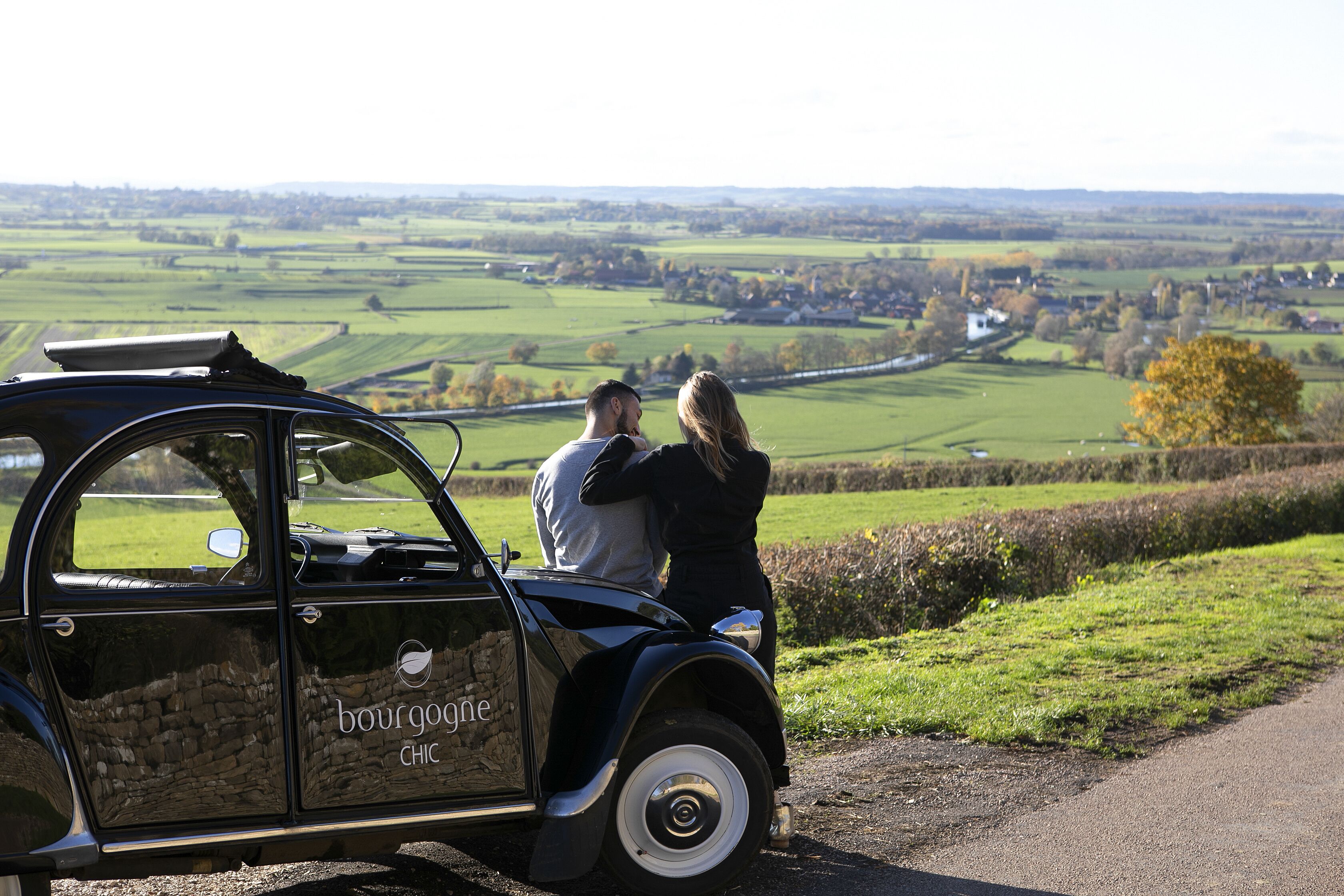 A vintage car parked on a rural road with a scenic view of fields and a small town in the distance.