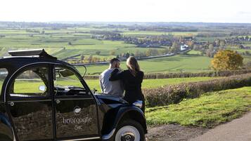 A vintage car parked on a rural road with a scenic view of fields and a small town in the distance.