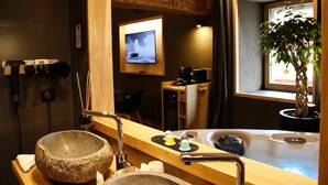 Une salle de bain moderne avec un lavabo en pierre, un meuble-lavabo en bois, un grand miroir, un téléviseur et une fenêtre agrémentée d’une plante en pot.