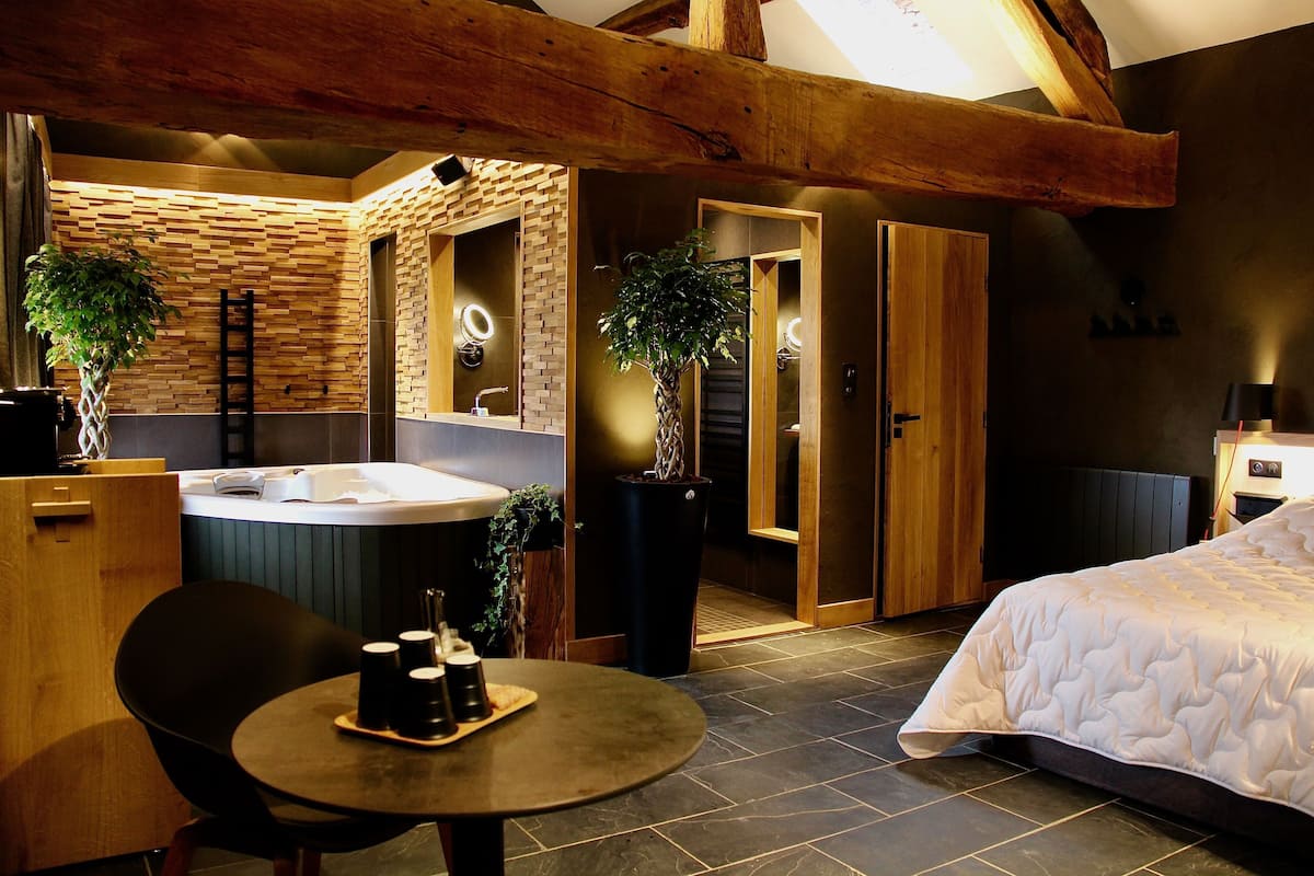 Une salle de bain moderne avec un grand miroir, un lavabo et un espace douche. On y trouve une table ronde avec une chaise et un lit recouvert d’une couette blanche.