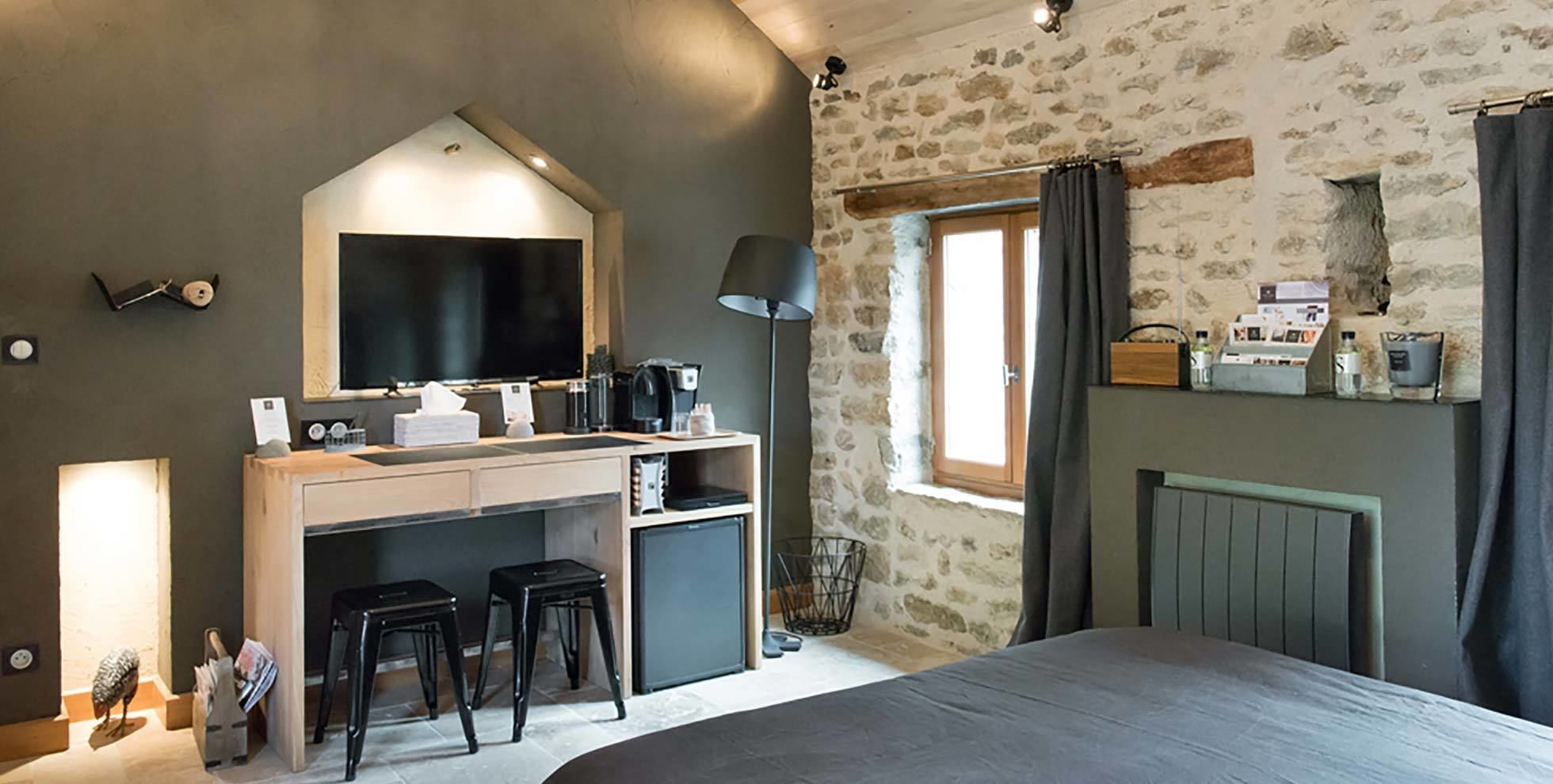 Une chambre avec un mur en pierre, un lit, un bureau avec un ordinateur, une lampe et une fenêtre avec des rideaux.