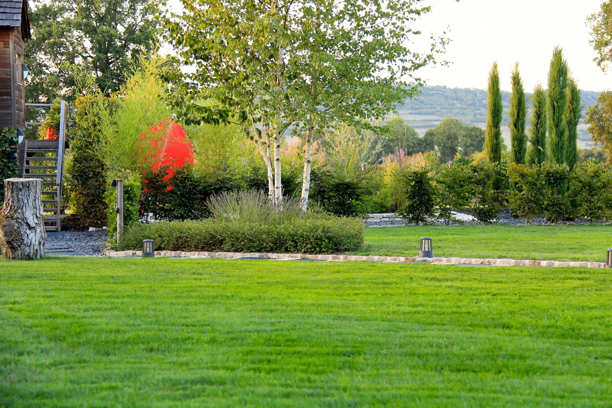 A well-maintained grassy lawn with a wooden fence, a stone pathway, and a variety of trees including a prominent red-leaved tree.