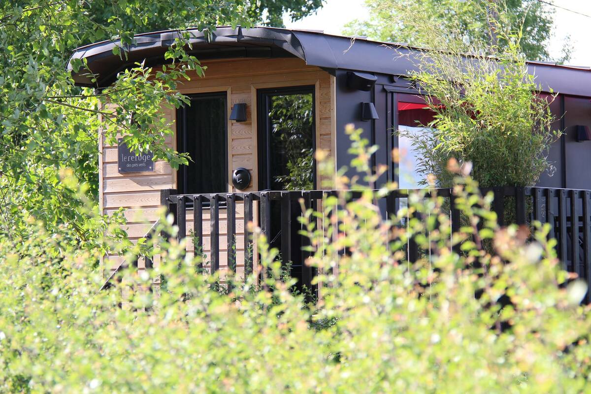 Une petite construction en bois, dotée d’une porte et de fenêtres, entourée de verdure.